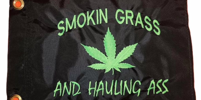 Smokin grass and hauling ass safety flag with dense embroidered green text and cannabis leaf on black nylon rectangle with solid brass grommets