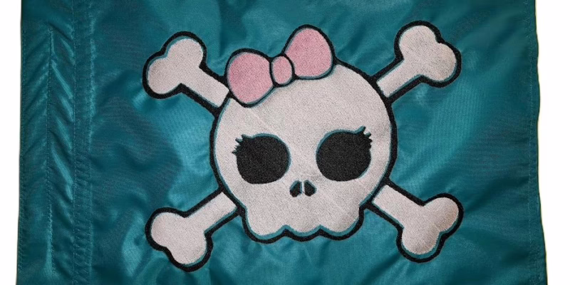 Alt Text: Teal rectangle safety whip flag featuring a dense embroidered white girly skull with a pink bow and eyelashes over crossbones, with a reinforced whip pole sleeve