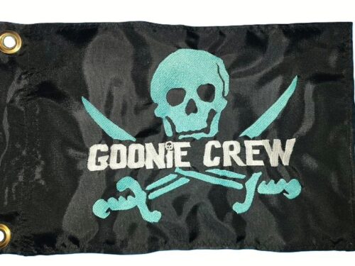 Black rectangle safety flag featuring embroidered teal skull and crossed cutlass swords with white GOONIE CREW text and two heavy-duty brass mounting grommets