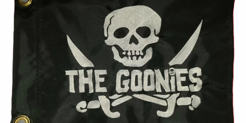Black rectangle safety flag featuring embroidered white skull and crossed cutlass swords with THE GOONIES block lettering and two heavy-duty brass mounting grommets