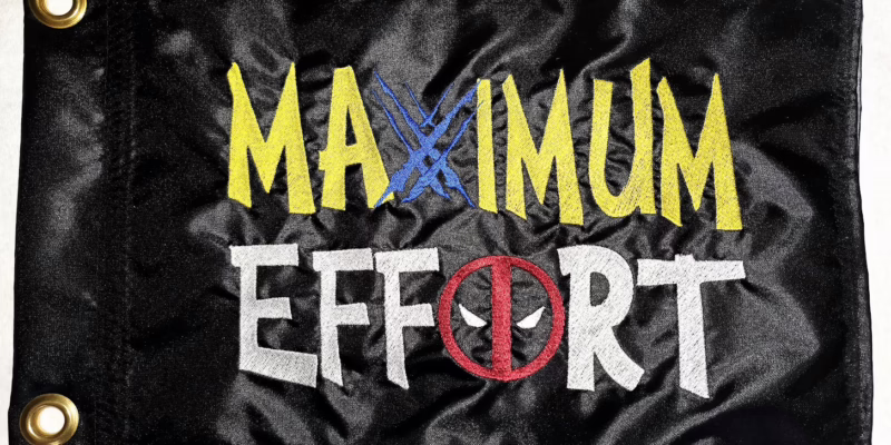Maximum Effort Deadpool and Wolverine safety flag with dense embroidered yellow and white lettering, blue claw marks, and masked logo on black nylon rectangle with brass grommets