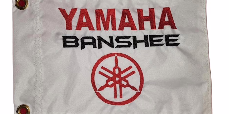 Yamaha Banshee safety flag with embroidered red Yamaha text, black Banshee lettering, and red tuning forks emblem on white nylon rectangle with brass grommets