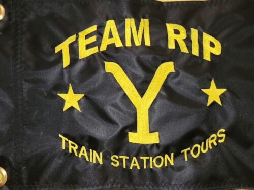 Team Rip Train Station Tours safety flag with dense embroidered yellow Yellowstone Y brand, stars, and text on black nylon rectangle with solid brass grommets