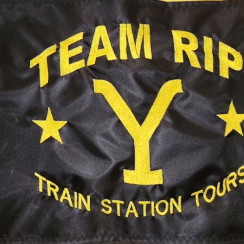 Team Rip Train Station Tours safety flag with dense embroidered yellow Yellowstone Y brand, stars, and text on black nylon rectangle with solid brass grommets