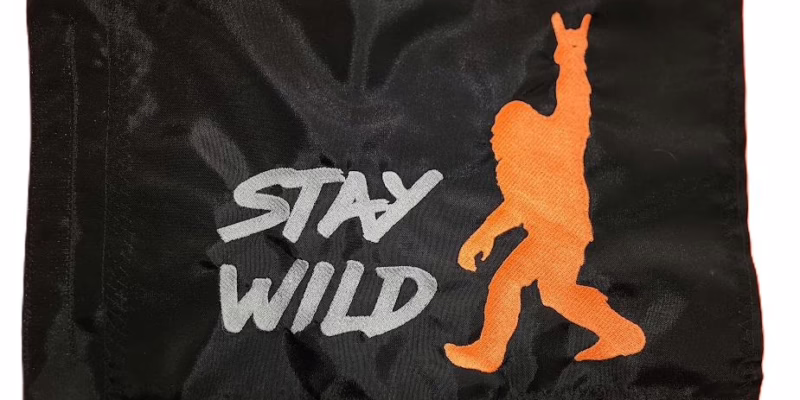 Stay Wild safety flag with embroidered orange Bigfoot throwing horns and silver lettering on black nylon rectangle with whip sleeve
