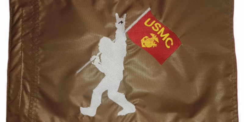Bigfoot USMC safety flag with embroidered silhouette holding Marine Corps banner on coyote tan nylon rectangle with whip sleeve
