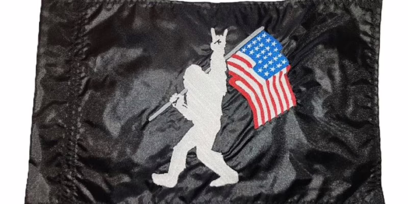 Rock On Bigfoot safety flag with embroidered white Sasquatch carrying American flag on black nylon rectangle with whip sleeve