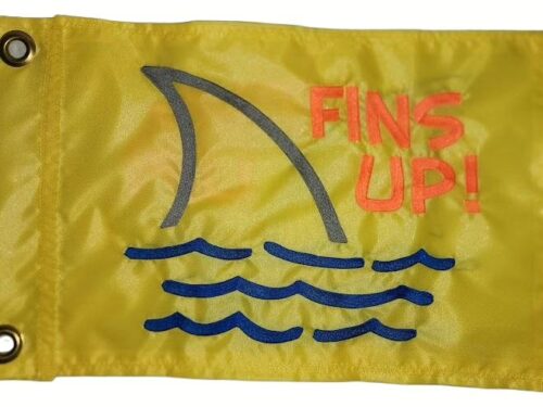 Fins Up safety flag with dense embroidered grey shark fin, blue water waves, and orange text on yellow nylon rectangle with solid brass grommets